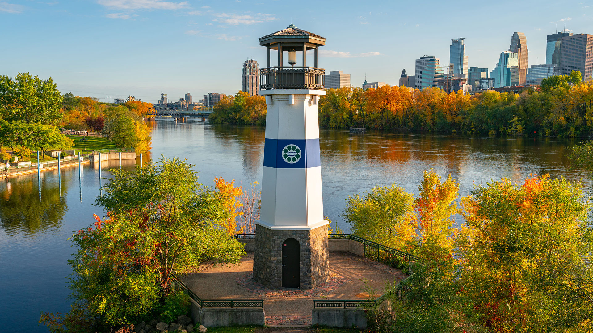 A tall lighthouse stands against a vibrant autumn backdrop, surrounded by colorful trees on a riverbank. City skyline visible in the distance under a clear sky.